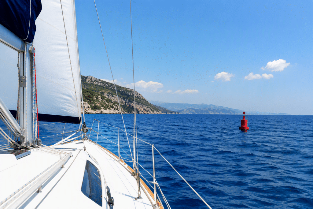 View from a sailboat with a white deck and rigging, blue sea and a rocky green coastline, a red buoy in the distance.